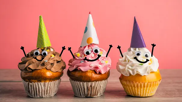 Drei Cupcakes mit bunten Cremehauben, Wackelaugen, lächelnden Gesichtern und Partyhüten stehen vor rosa Hintergrund. - Foto: sdominick/iStock Drei Cupcakes mit bunten Cremehauben, Wackelaugen, lächelnden Gesichtern und Partyhüten stehen vor rosa Hintergrund. - Foto: sdominick/iStock