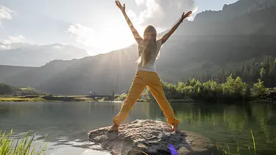 Junge Frau rümpft sich bei Sonnenaufgang auf Felsen über Alpensee - Foto: Mystockimages / iStock