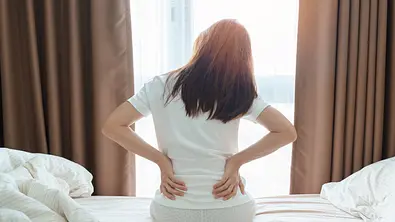 Frau sitzt auf Ihrem Bett und hält mit beiden Händen ihren unteren Rücken - Foto: Panuwat Dangsungnoen / iStock
