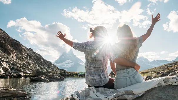 Zwei Menschen sitzen eng umschlungen an einem Bergsee und strecken die Arme in die Höhe. Die Sonne strahlt über den Gipfeln. - Foto: Mystockimages/iStock Zwei Menschen sitzen eng umschlungen an einem Bergsee und strecken die Arme in die Höhe. Die Sonne strahlt über den Gipfeln. - Foto: Mystockimages/iStock