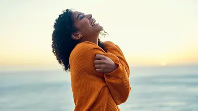 Eine Frau mit lockigem Haar und orangefarbenem Pullover lacht mit verschränkten Armen vor dem Meer in der Abendsonne. - Foto: pixdeluxe/iStock Eine Frau mit lockigem Haar und orangefarbenem Pullover lacht mit verschränkten Armen vor dem Meer in der Abendsonne. - Foto: pixdeluxe/iStock