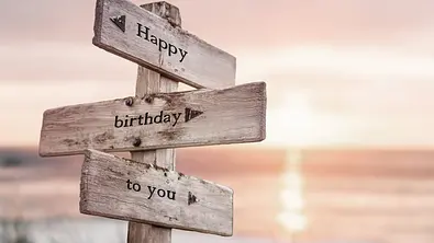 Happy Birthday to you auf einem hölzernen Wegweiser am Strand bei Sonnenuntergang. - Foto: Jon Anders Wiken / AdobeStock