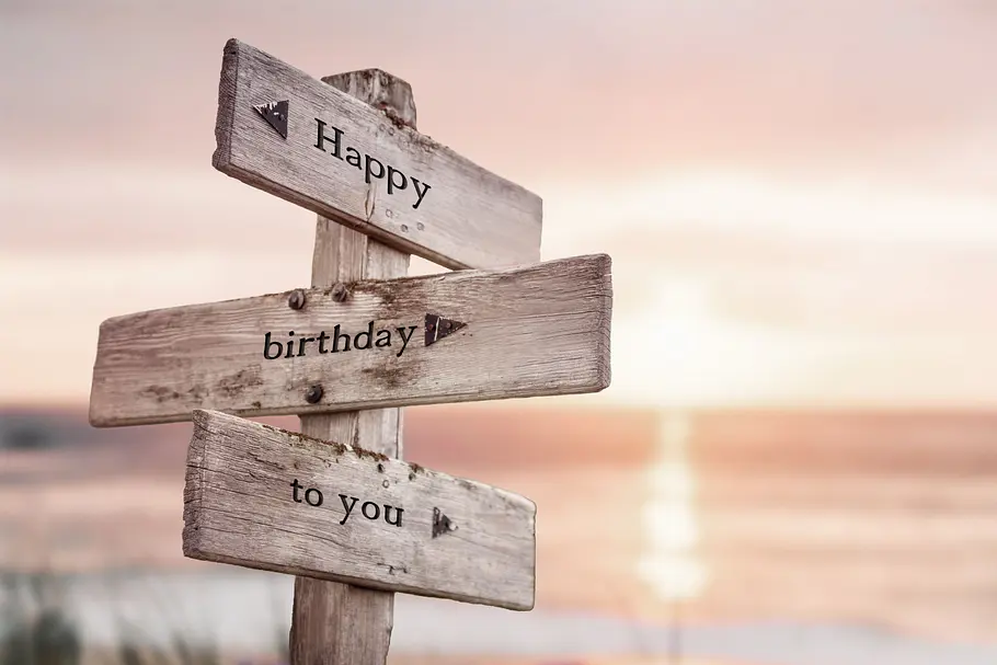Happy Birthday to you auf einem hölzernen Wegweiser am Strand bei Sonnenuntergang.