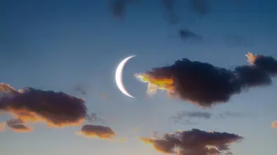 Eine helle Mondsichel leuchtet am blauen Himmel zwischen dunklen Wolken mit orange leuchtenden Rändern. - Foto: Nazarii Neshcherenskyi/iStock