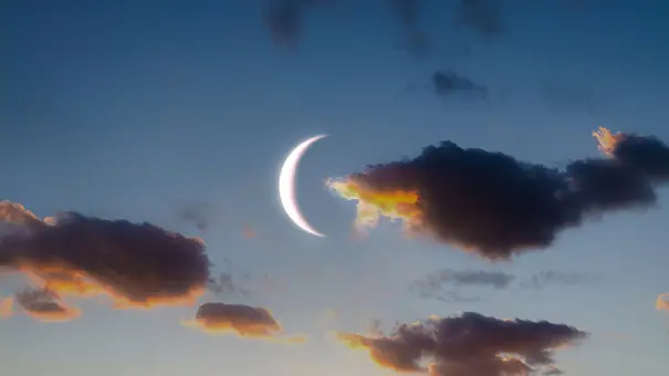 Eine helle Mondsichel leuchtet am blauen Himmel zwischen dunklen Wolken mit orange leuchtenden Rändern. - Foto: Nazarii Neshcherenskyi/iStock Eine helle Mondsichel leuchtet am blauen Himmel zwischen dunklen Wolken mit orange leuchtenden Rändern. - Foto: Nazarii Neshcherenskyi/iStock