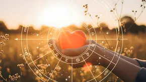 Sternzeichenkreis umringt eine Hand, die ein rotes Herz vor einem Feld im Sommer hält - Foto: Collage mit AdobeStock (Artfinity / Назарій) und Astrowoche.de