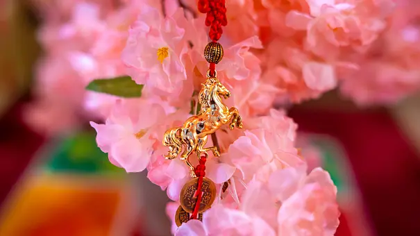 Auf dem Foto ist ein goldenes Pferde‑Amulett an einer roten Schnur vor rosa Blüten zu sehen, das symbolisch für das chinesische Jahr des Feuerpferdes steht. - Foto: yuki-ramen1025/iStock Auf dem Foto ist ein goldenes Pferde‑Amulett an einer roten Schnur vor rosa Blüten zu sehen, das symbolisch für das chinesische Jahr des Feuerpferdes steht. - Foto: yuki-ramen1025/iStock