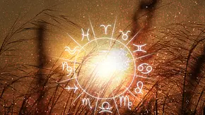Das Bild zeigt eine Grafik des astrologischen Tierkreises vor einem verschwommenen Hintergrund eines Feldes in der Natur, das gold-glitzernd schimmert. - Foto: Liudmila Chernetska/iStock