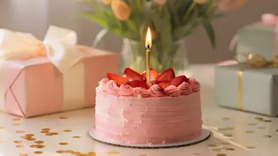 Eine rosa Torte mit Erdbeeren und einer brennenden Kerze steht auf einem festlich dekorierten Tisch neben Geschenken und einem Tulpenstrauß. - Foto: AdobeFirefly (generiert mit KI)