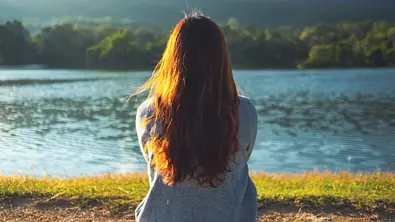 Frau in einem grauen Pullover sitzt an einem See vor einer Berglandschaft - Foto: Farknot Architect / AdobeStock Frau in einem grauen Pullover sitzt an einem See vor einer Berglandschaft - Foto: Farknot Architect / AdobeStock