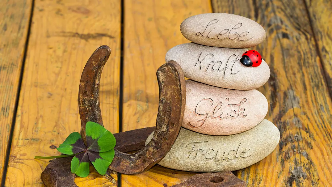 Beschriftete Steine mit „Liebe“, „Kraft“, „Glück“ und „Freude“, zwei Hufeisen, ein Kleeblatt und ein Deko-Marienkäfer liegen auf Holz. - Foto: Sonja Calovini/AdobeStock