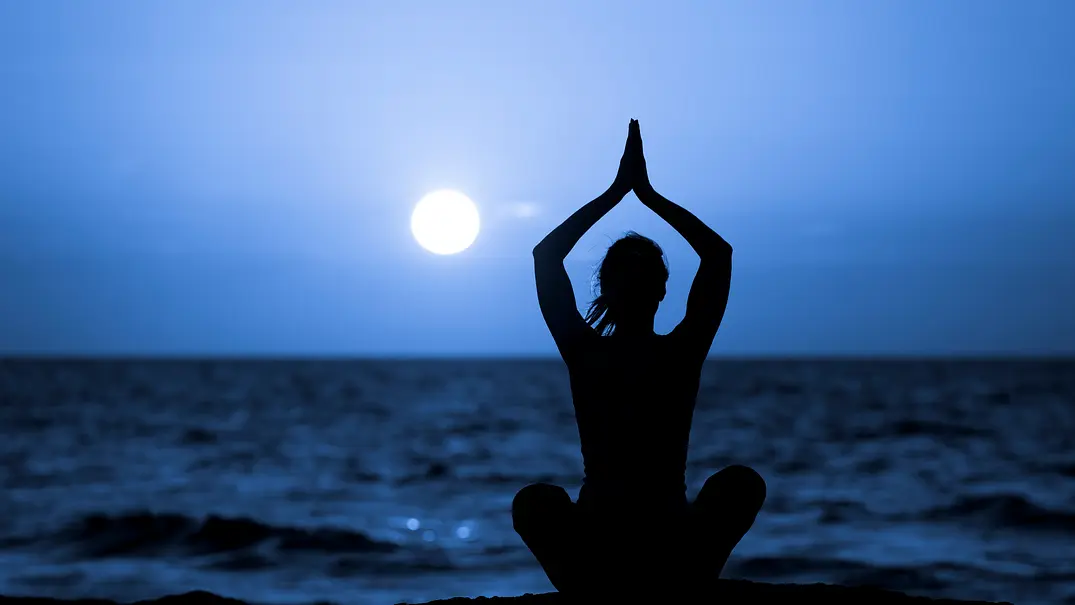 Eine Person sitzt abends als Silhouette am Meer und hält die Hände über dem Kopf zusammen. Am dunklen Himmel leuchtet der Mond. - Foto: AnnaRise/iStock Eine Person sitzt abends als Silhouette am Meer und hält die Hände über dem Kopf zusammen. Am dunklen Himmel leuchtet der Mond. - Foto: AnnaRise/iStock