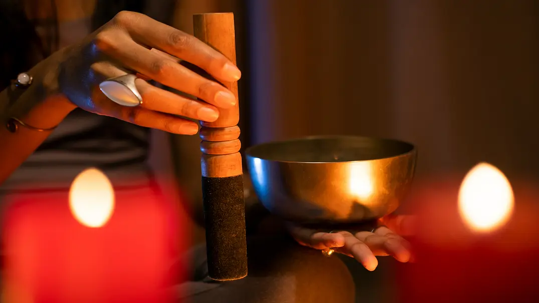 Eine Person hält eine goldfarbene Klangschale und einen Holzschlägel. Zwei rote Kerzen leuchten unscharf im Vordergrund. - Foto: D-Keine/iStock