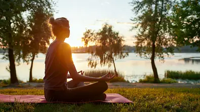 Frau sitzt im Lotussitz an einem See in der Sonne - Foto: Svitlana / AdobeStock