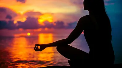 Frau sitzt meditierend vor einem leuchtenden Sonnenuntergang über dem Meer - Foto: guruXOX / AdobeStock