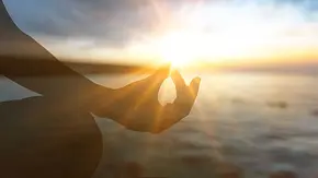 Frau hält Hand in Meditationspose vor der Sonne über dem Meer - Foto: BillionPhotos.com / AdobeStock Frau hält Hand in Meditationspose vor der Sonne über dem Meer - Foto: BillionPhotos.com / AdobeStock