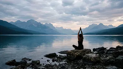 Eine Person sitzt mit erhobenen Armen auf einem Felsen am See. Im Hintergrund liegen Berge unter einem bewölkten Himmel. - Foto: SALMAAKTER/AdobeStock, KI generiert