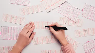 Eine Person füllt mehrere Lottoscheine auf einem Tisch aus und setzt mit einem Stift Kreuzchen auf den Feldern. - Foto: LIGHTFIELD STUDIOS / AdobeStock