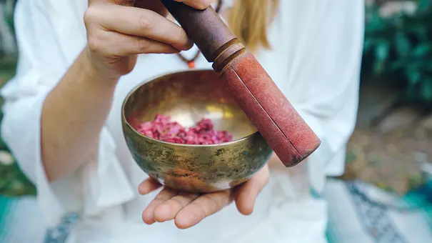 Eine Person in Weiß hält eine messingfarbene Klangschale mit rosa Blütenblättern und einen rot gepolsterten Holzschlägel. - Foto: Juanmarcos/AdobeStock