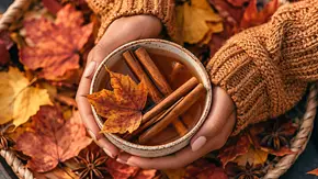 Hände in einem braunen Strickpullover halten eine Tasse mit Zimtstangen und einem Ahornblatt, umgeben von Herbstlaub und Sternanis. - Foto:  Raul/AdobeStock, KI generiert